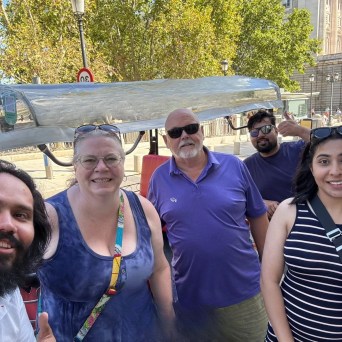 Five people smiling outdoors with trees and a building in the background.