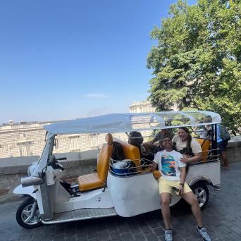 Group sitting in white tuk-tuk on cobblestone path near trees and building.