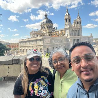 Three people smiling in front of a large cathedral on a sunny day.
