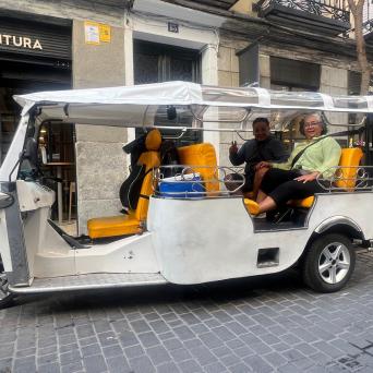 Two people sitting in a white tuk-tuk with yellow seats on a city street.