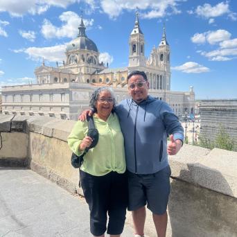 Two people smiling in front of a historic building with blue sky and clouds.