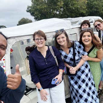 Group of people smiling and posing outdoors near a small white vehicle with trees in the background.