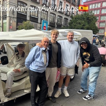 Five people smiling near a decorated tuk-tuk in a city square with outdoor seating.