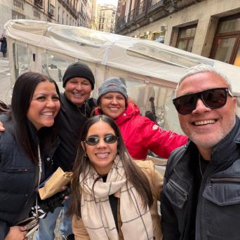 Group of five people smiling and posing outdoors with buildings in the background.