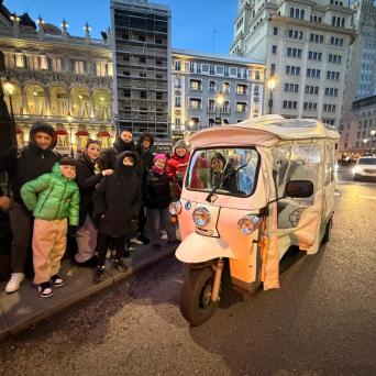 Group of people pose by a tuk-tuk on a city street with illuminated buildings in the evening.