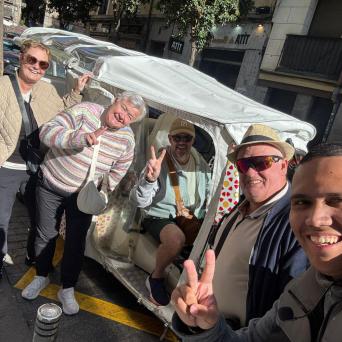 Group of five people posing with peace signs in front of a covered rickshaw on a sunny street.