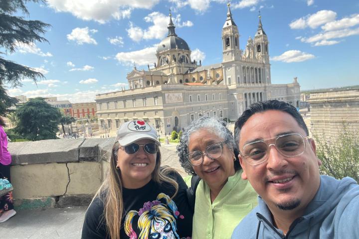 Three people smiling in front of a large cathedral on a sunny day.