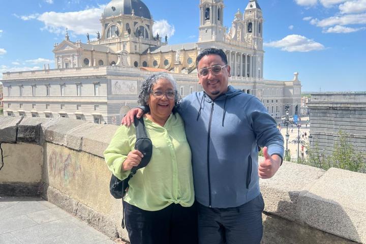 Two people smiling in front of a historic building with blue sky and clouds.