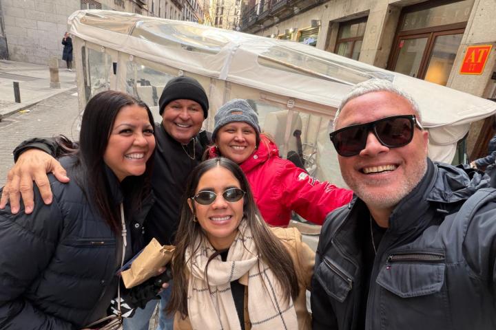Group of five people smiling and posing outdoors with buildings in the background.