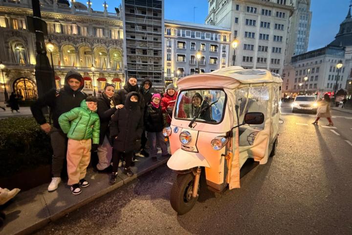 Group of people pose by a tuk-tuk on a city street with illuminated buildings in the evening.